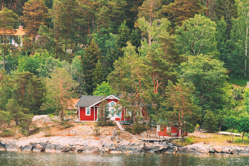 Sweden. Beautiful Red ASwedish Wooden Log Cabins Houses On Rocky Island Coast In Summer. Lake Or River Landscape